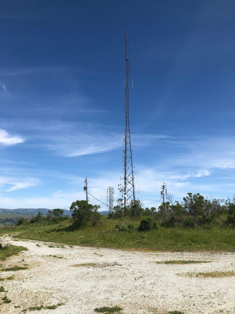 The top of Coyote Peak: a white gravel path surrounds      a small patch of grass, atop which sits a huge antenna tower alongside      more transmission towers and poles, in the background is a blue sky and a line of hills in the distance.