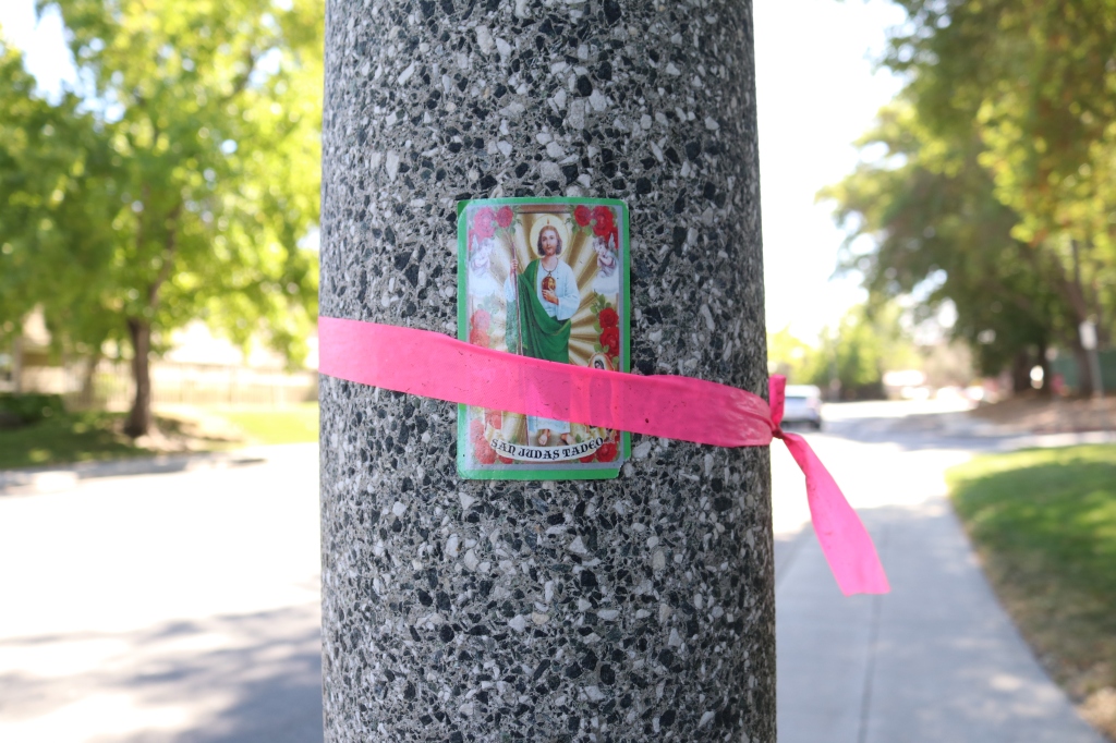 A San Judas Tadeo prayer card is tied to a black and white speckled light pole with a thin piece of pink plastic.  The background is a sunny, tree lined street with a car driving on the righthand side.  On the prayer card San Judas Tadeo is weiaring a green robe over a white tunic.  he is holding something that is not 100% visible.  He is surrounded by red roses, and gold and yellow light is emanating from him. 